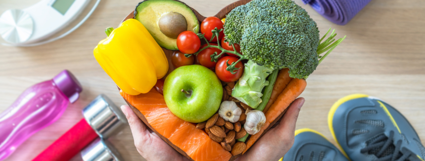 Heart dish with healthy foods, weights, scale, and sports shoes in fitness center.