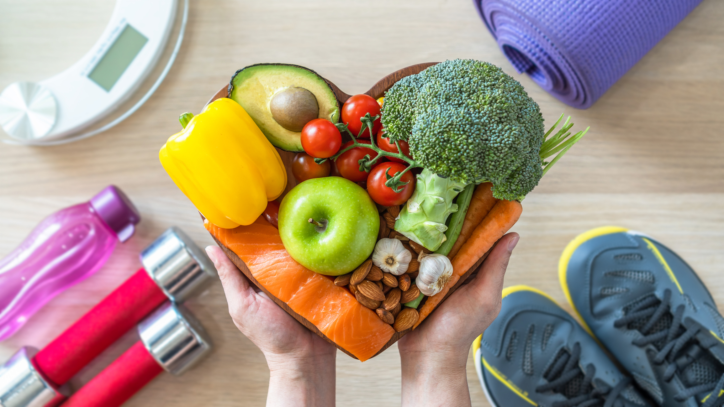 Heart dish with healthy foods, weights, scale, and sports shoes in fitness center.