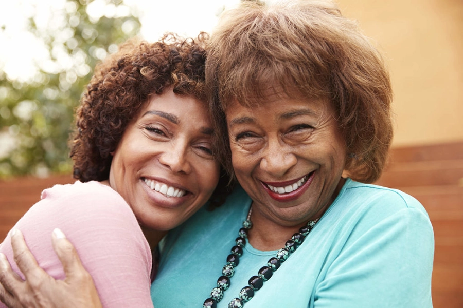Two women, one younger and one older, smiling and hugging each other warmly outdoors. The younger woman is wearing a pink top, and the older woman is wearing a light blue top with a beaded necklace.